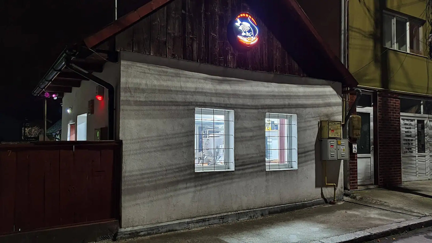 Panoul de neon iluminat deasupra intrării clădirii prezintă un design circular strălucitor cu o schemă de culori portocaliu și albastru, reprezentând o figură stilizată de animal.